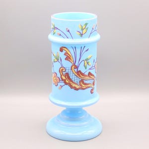Puede incluir: Un jarrón de cristal azul claro con base de pedestal y cuerpo cilíndrico. El jarrón está decorado con diseños florales y de volutas en dorado, naranja y morado. El borde superior presenta una banda de hojas y bayas.