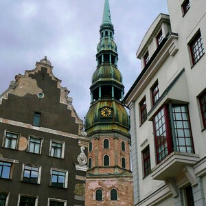 May include: A tall, green and brown church steeple with a clock face and a pointed roof. The church is surrounded by older buildings with a gray sky in the background.