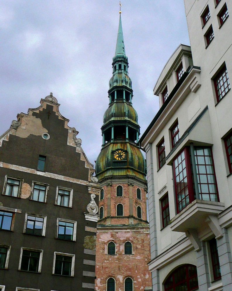 May include: A tall, green and brown church steeple with a clock face and a pointed roof. The church is surrounded by older buildings with a gray sky in the background.