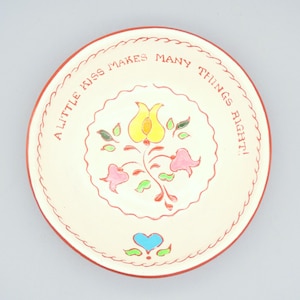 May include: A cream-colored ceramic plate with a red rim and the text "A LITTLE KISS MAKES MANY THINGS RIGHT!" in red. The plate features a floral design with yellow, pink, and green accents.