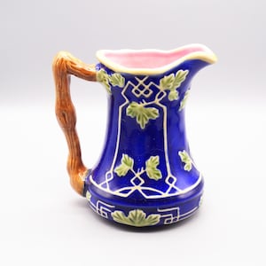 May include: A blue ceramic pitcher with a brown handle and a pink interior. The pitcher is decorated with green leaves and white geometric patterns. A decorative piece for the kitchen or dining room.