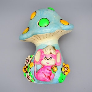 Pode incluir: Uma figura de cogumelo de cerâmica caprichosa com uma tampa azul clara adornada com manchas amarelas e verdes. Um rato rosa com orelhas grandes está aninhado na base, rodeado por flores coloridas. A figura é pintada com cores brilhantes e alegres.