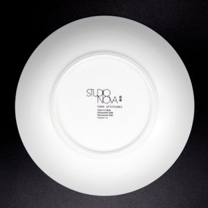 May include: White ceramic plate with a black logo that reads "Studio Nova" and "Y2005 Attitudes" with the text "Oven to Table Dishwasher Safe Microwave Safe Thailand - 0"