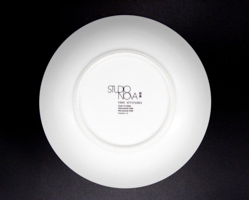 May include: White ceramic plate with a black logo that reads "Studio Nova" and "Y2005 Attitudes" with the text "Oven to Table Dishwasher Safe Microwave Safe Thailand - 0"