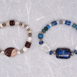 May include: Two beaded bracelets with different coloured stones. One bracelet has brown and white stones with a large brown and white stone in the centre. The other bracelet has blue and white stones with a large blue stone in the centre.