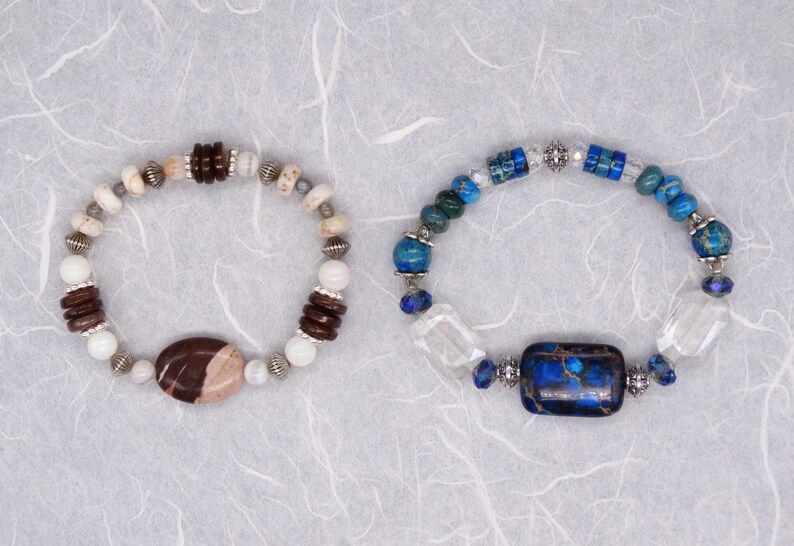 May include: Two beaded bracelets with different coloured stones. One bracelet has brown and white stones with a large brown and white stone in the centre. The other bracelet has blue and white stones with a large blue stone in the centre.