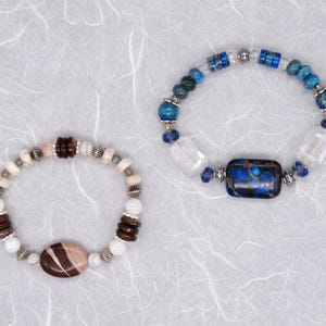 May include: Two bracelets featuring a variety of coloured stones and metal accents. One bracelet has blue, white and brown stones, while the other has white, brown and silver stones.