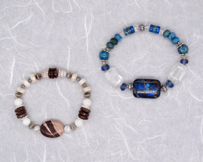 May include: Two bracelets featuring a variety of coloured stones and metal accents. One bracelet has blue, white and brown stones, while the other has white, brown and silver stones.