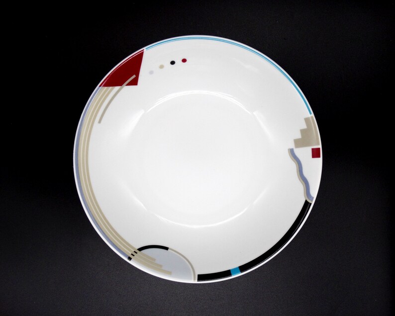 May include: A white ceramic bowl with a geometric design in shades of blue, red, and gray. The bowl has a wide rim and a shallow depth.