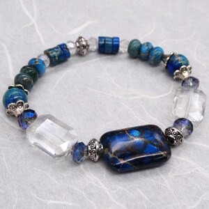 May include: A blue and white beaded bracelet with a large rectangular blue and brown jasper stone. The bracelet also features clear crystal beads and silver accents.