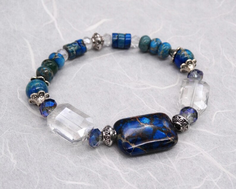 May include: A blue and white beaded bracelet with a large rectangular blue and brown jasper stone. The bracelet also features clear crystal beads and silver accents.
