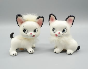 May include: Two white ceramic kittens with black ears and tails. The kittens have pink noses and black eyes with white pupils. They are both sticking their tongues out.