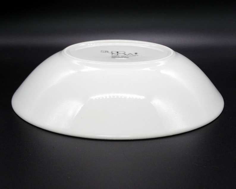 May include: White ceramic serving bowl with a slightly curved rim. The bottom of the bowl has the text "Studio Nova" printed in black.