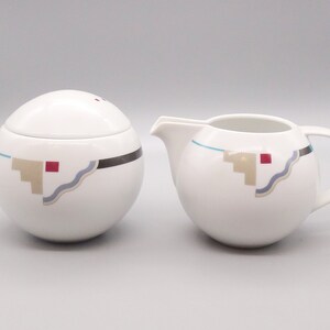 May include: A white ceramic sugar bowl and creamer set with a geometric design in shades of brown, blue, and red. The design features a wavy line, a series of steps, and a square.
