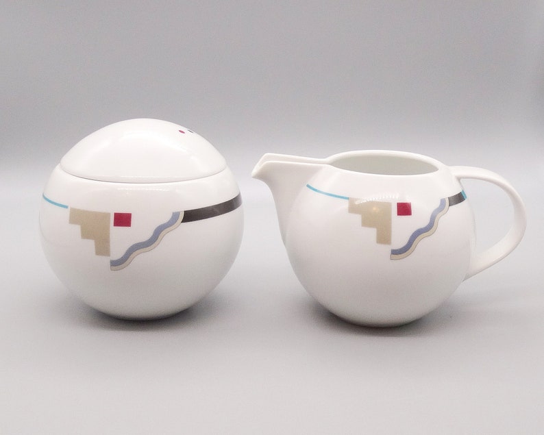 May include: A white ceramic sugar bowl and creamer set with a geometric design in shades of brown, blue, and red. The design features a wavy line, a series of steps, and a square.