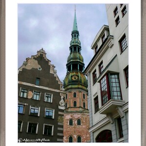May include: A tall, green-roofed church with a clock tower stands next to a white building with a large window. The church has a pointed spire and a brick facade. The white building has a flat roof and a row of windows.