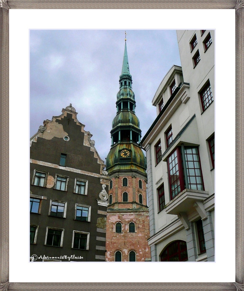 May include: A tall, green-roofed church with a clock tower stands next to a white building with a large window. The church has a pointed spire and a brick facade. The white building has a flat roof and a row of windows.