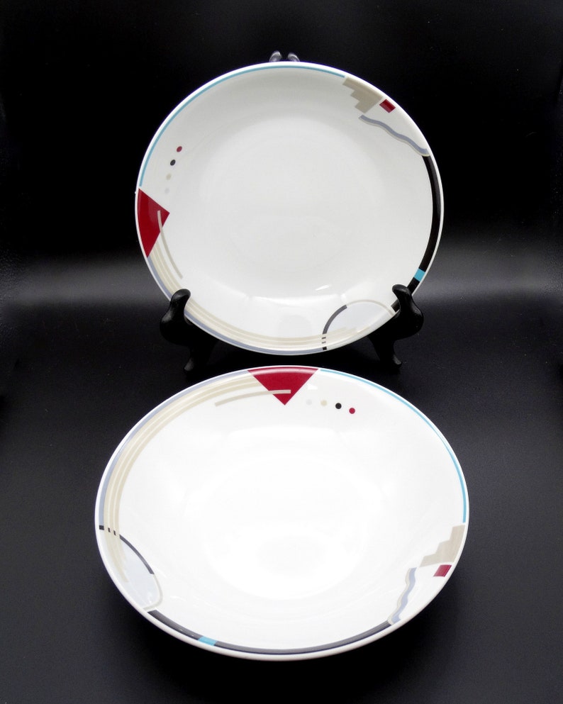 May include: Two white ceramic bowls with a geometric design in shades of red, blue, gray, and black. The bowls have a white interior and a rim with a blue and gray stripe.