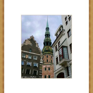May include: A tall, green-roofed church tower with a clock face stands in the middle of a narrow street in a European city. The church is flanked by two older buildings with brown brick facades and many windows.