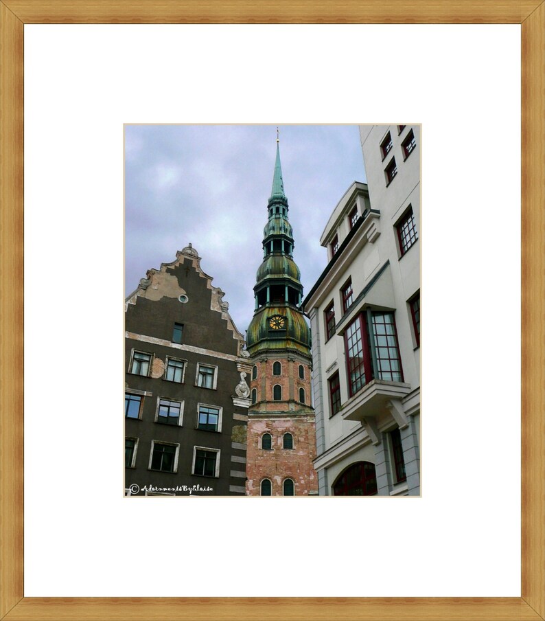 May include: A tall, green-roofed church tower with a clock face stands in the middle of a narrow street in a European city. The church is flanked by two older buildings with brown brick facades and many windows.