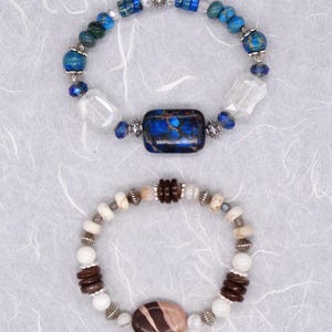 May include: Two bracelets featuring different coloured stones. The top bracelet has blue and green stones with a large blue and brown stone in the centre. The bottom bracelet has white and brown stones with a large brown and pink stone in the centre.