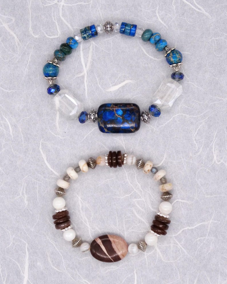 May include: Two bracelets featuring different coloured stones. The top bracelet has blue and green stones with a large blue and brown stone in the centre. The bottom bracelet has white and brown stones with a large brown and pink stone in the centre.