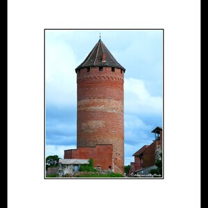 Op de afbeelding: Een hoge, rode bakstenen toren met een puntdak en een klein raam bovenaan. De toren is omgeven door een stenen muur en een grasveld.