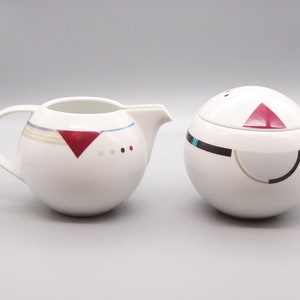 May include: A white ceramic creamer and sugar bowl set with geometric designs in red, black, and blue. The creamer has a handle and a spout. The sugar bowl has a lid.