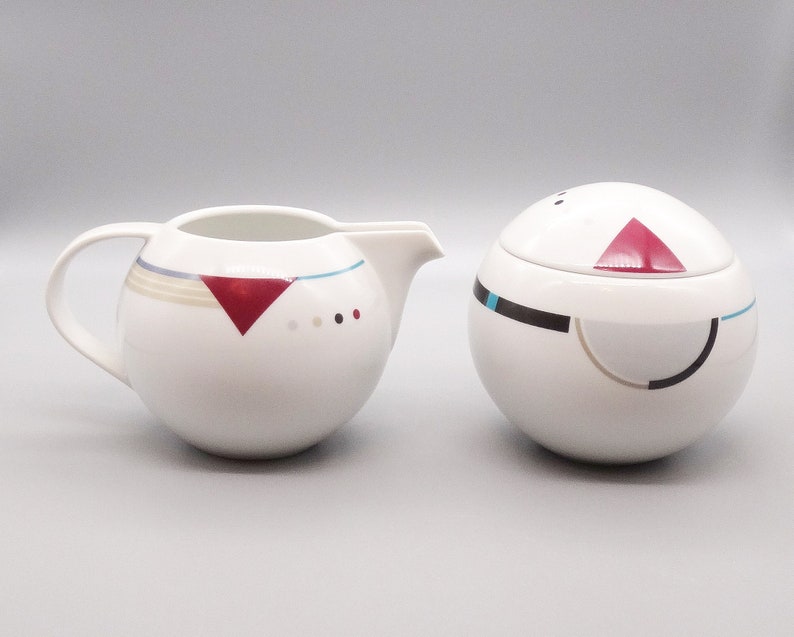 May include: A white ceramic creamer and sugar bowl set with geometric designs in red, black, and blue. The creamer has a handle and a spout. The sugar bowl has a lid.