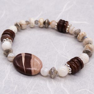 May include: A brown and white beaded bracelet featuring a large oval-shaped brown and white stone at its centre. The bracelet is crafted with a variety of bead sizes and shapes, including round, oval, and flat.