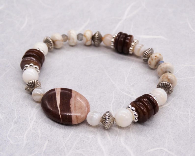 May include: A brown and white beaded bracelet featuring a large oval-shaped brown and white stone at its centre. The bracelet is crafted with a variety of bead sizes and shapes, including round, oval, and flat.