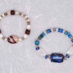 May include: Two bracelets featuring different coloured stones and metal accents. One bracelet has brown and white stones with a brown oval-shaped stone in the centre. The other bracelet has blue and white stones with a blue rectangular stone in the centre.