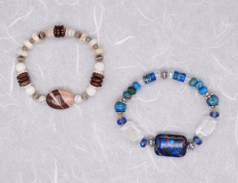 May include: Two bracelets featuring different coloured stones and metal accents. One bracelet has brown and white stones with a brown oval-shaped stone in the centre. The other bracelet has blue and white stones with a blue rectangular stone in the centre.