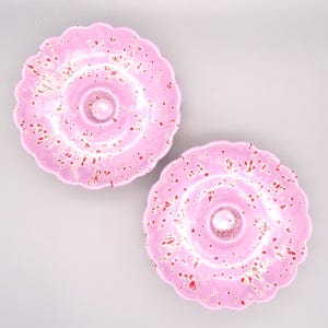 May include: Two round ceramic ring dishes with a scalloped edge, featuring a glossy light pink glaze with a speckled pattern of dark red and white paint splatters. Each dish has a small, raised central post for holding rings or jewelry.