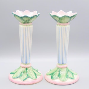 Puede incluir: Par de candelabros decorativos de cerámica. Cada uno tiene una base y una parte superior rosas, con detalles de hojas verdes. La columna central es blanca con rayas verticales azules. La parte superior tiene un diseño rosa en forma de flor.