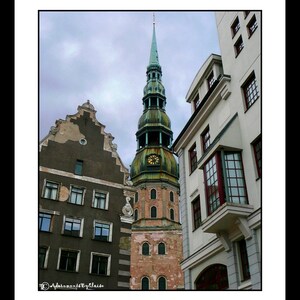 May include: A tall, green-roofed church with a clock tower stands in the middle of the image. The church is surrounded by older buildings with red tile roofs. The sky is cloudy and gray.