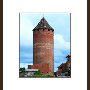 Op de afbeelding: Een hoge, rode bakstenen toren met een puntig dak staat tegen een bewolkte lucht. De toren heeft een reeks kleine ramen en een decoratief baksteenpatroon rond de top.