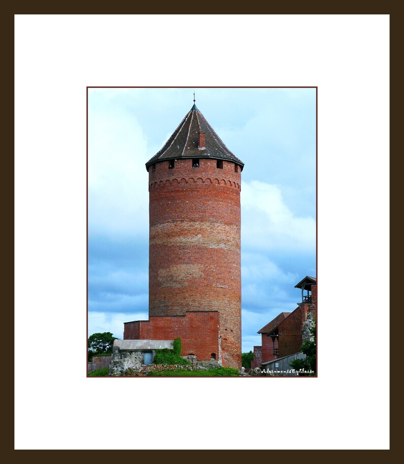 Op de afbeelding: Een hoge, rode bakstenen toren met een puntig dak staat tegen een bewolkte lucht. De toren heeft een reeks kleine ramen en een decoratief baksteenpatroon rond de top.
