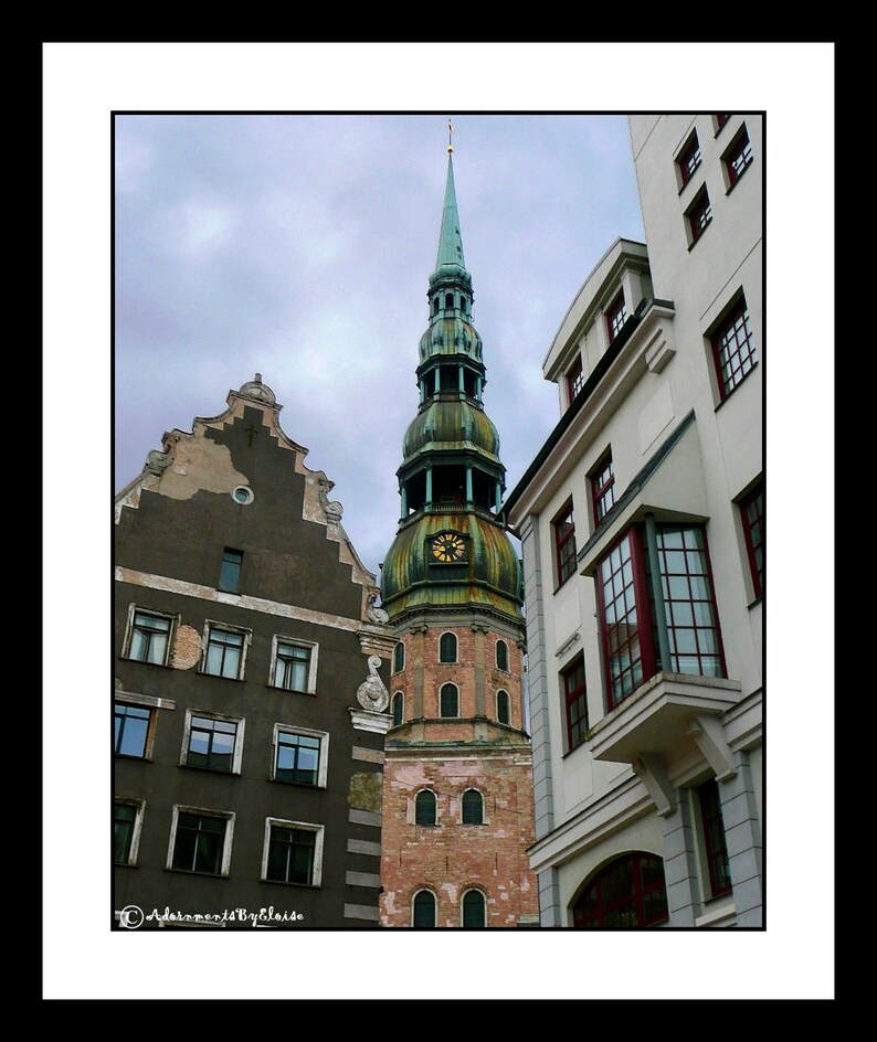 May include: A tall, green-roofed church with a clock tower stands in the middle of the image. The church is surrounded by older buildings with red tile roofs. The sky is cloudy and gray.