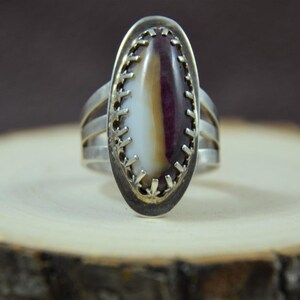 May include: A silver ring with a purple, white, and brown oval stone set in a decorative silver bezel. The ring has three bands.