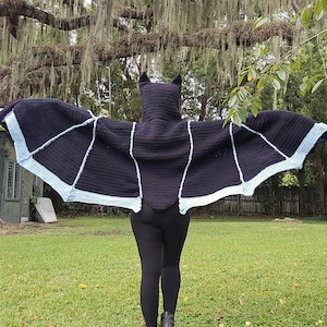 May include: A black crocheted bat wing garment with a hood featuring pointed ears. The wingspan is wide, showcasing a solid black main body with light blue trim along the bottom edges of the wings. The garment is worn over black leggings.