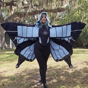 May include: A crochet butterfly wing shawl featuring a hood, designed with a color palette of black and light blue. The garment displays a large, symmetrical wing pattern that extends outward, crafted with detailed stitching to mimic butterfly wings.