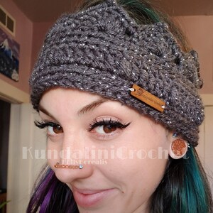 May include: A gray crocheted headband with silver glitter, featuring a crown design and a brown leather tag with the text "Kundalini Crochet" in white lettering. The headband is worn by a person with purple and teal hair, and they are wearing a nose ring with multiple beads.
