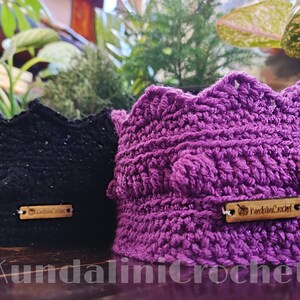 May include: Two crocheted plant pot covers, one black and one purple, with wooden tags that say "Kundalini Crochet" and "Lodie's Craft".