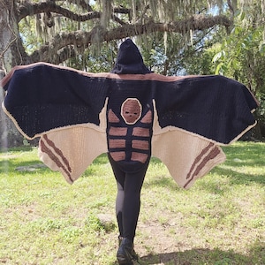 May include: A handmade crochet bat wing hooded cape in navy blue, brown, and beige. The cape features a bat-like design with a hood and a detailed face on the back. The wings are spread out, showcasing the intricate crochet work.