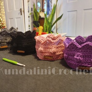 May include: Four crocheted crown-shaped headbands in different colors: gray, black, pink, and purple. The headbands are made with a textured stitch and have a small tag attached to each.