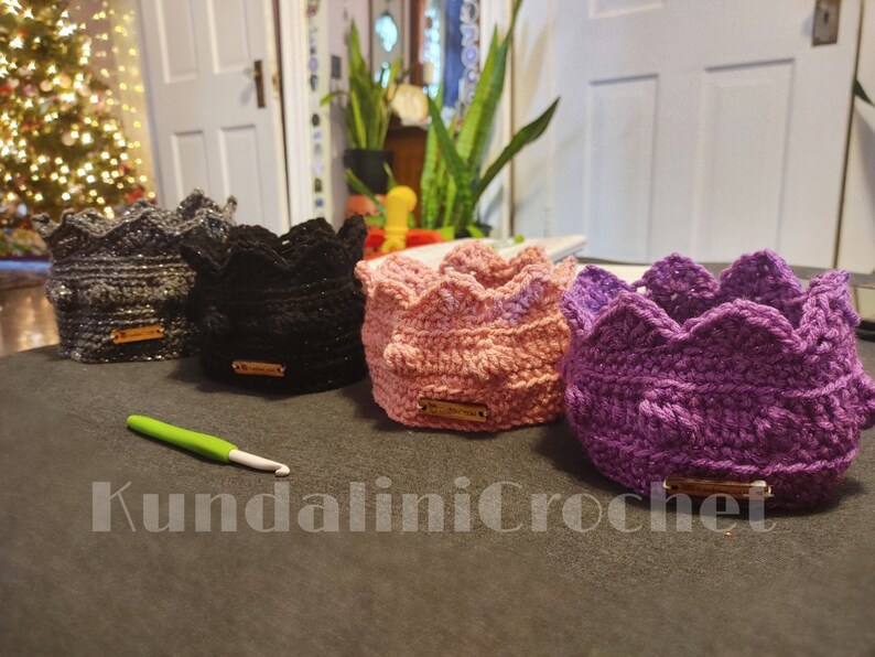 May include: Four crocheted crown-shaped headbands in different colors: gray, black, pink, and purple. The headbands are made with a textured stitch and have a small tag attached to each.
