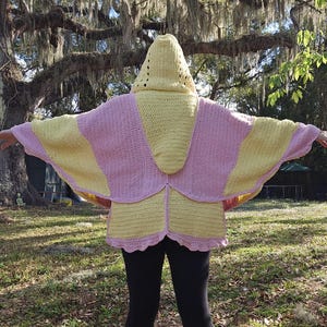 May include: A handmade crochet hooded cape in shades of yellow and pink. The cape features a hood with eyelet details and a scalloped hemline. The wings are spread out, showcasing the color-blocked design.