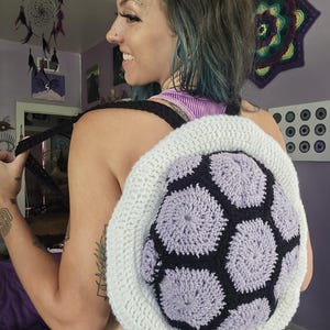 May include: A handmade crochet backpack with a unique design. The backpack features a central section with a pattern of lavender and black hexagons, framed by a white border. The straps are black.
