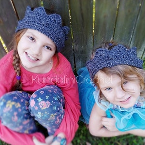 May include: Two young girls wearing gray crocheted crown headbands. The girl on the left is wearing a pink sweater and blue jeans with a floral pattern. The girl on the right is wearing a blue tulle dress.